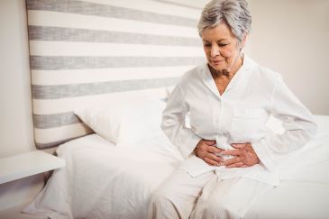 Senior woman suffering from stomach ache sitting on bed in bedroom