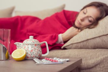 Sick woman covered with a blanket lying in bed with high fever and a flu, resting. Teapot, pills and lemon on the table, focus on the teapot