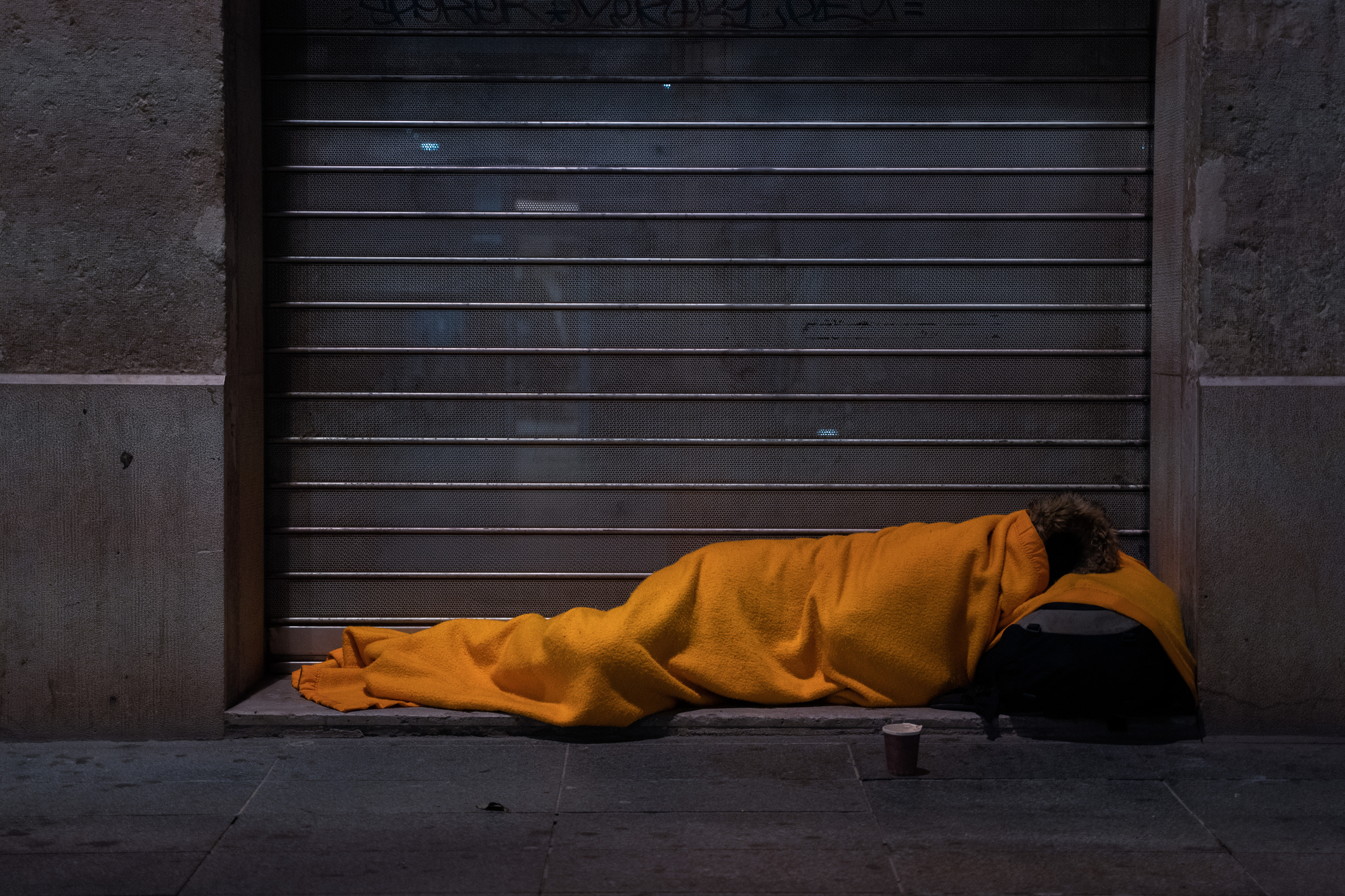 A homeless person sleeps on the street under a blanket in front of a storefront.